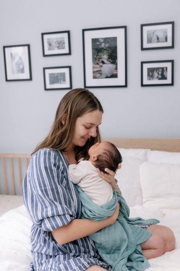 A mother sits on a bed and smiles down at a newborn wrapped in a teal blanket.