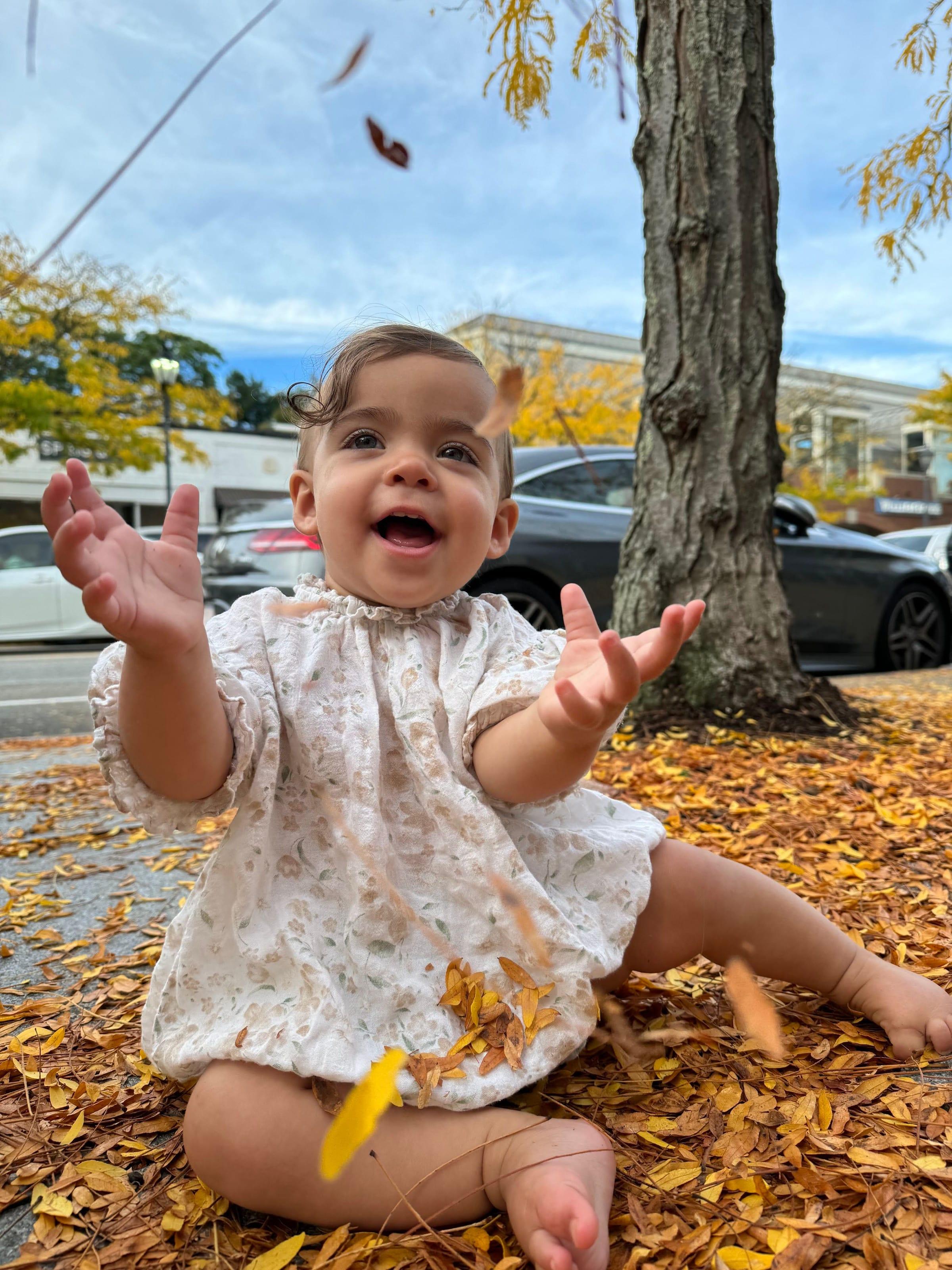A smiling baby sits among fallen autumn leaves with arms raised as a leaf floats by.