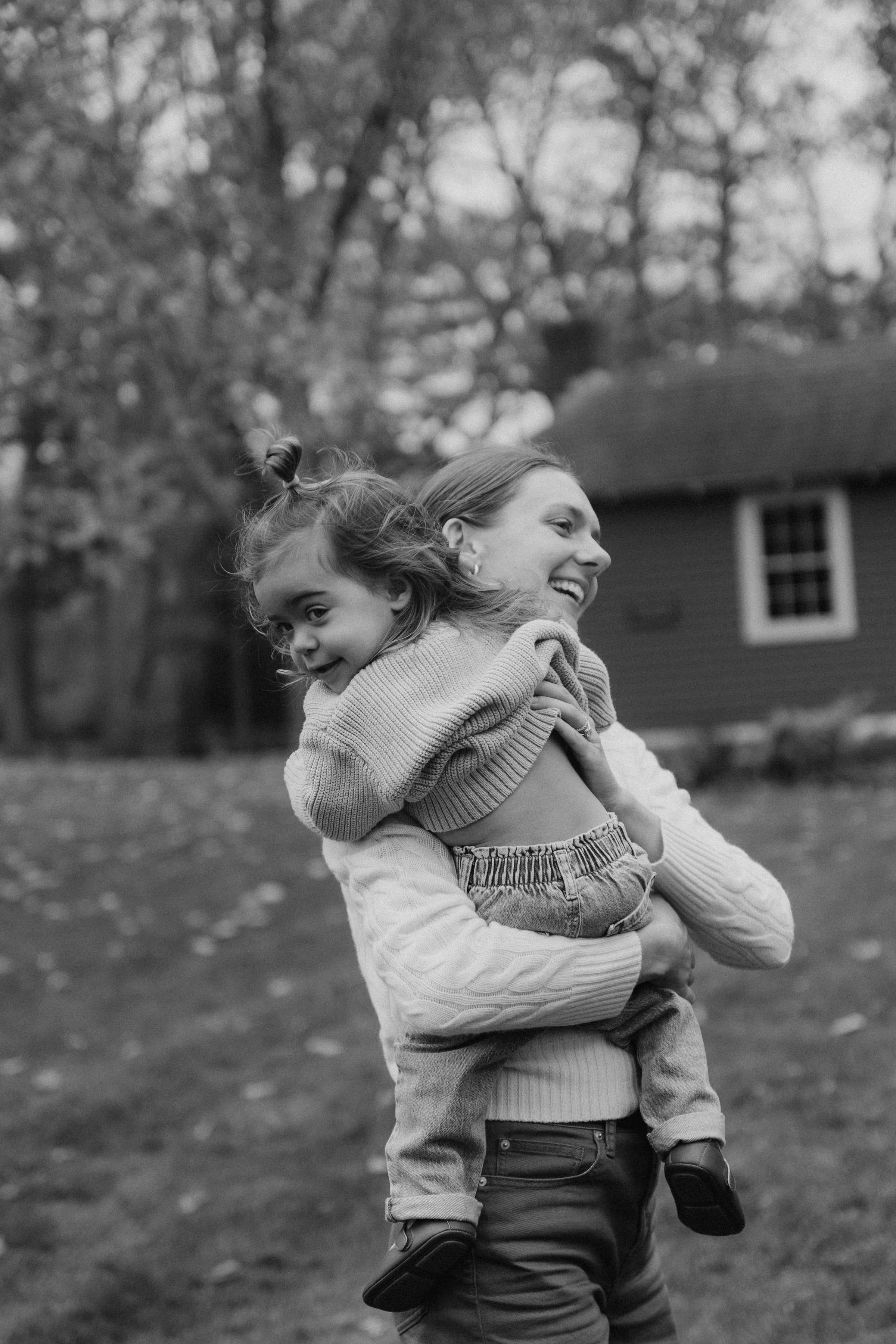 A smiling mother carries a toddler on her back outdoors.