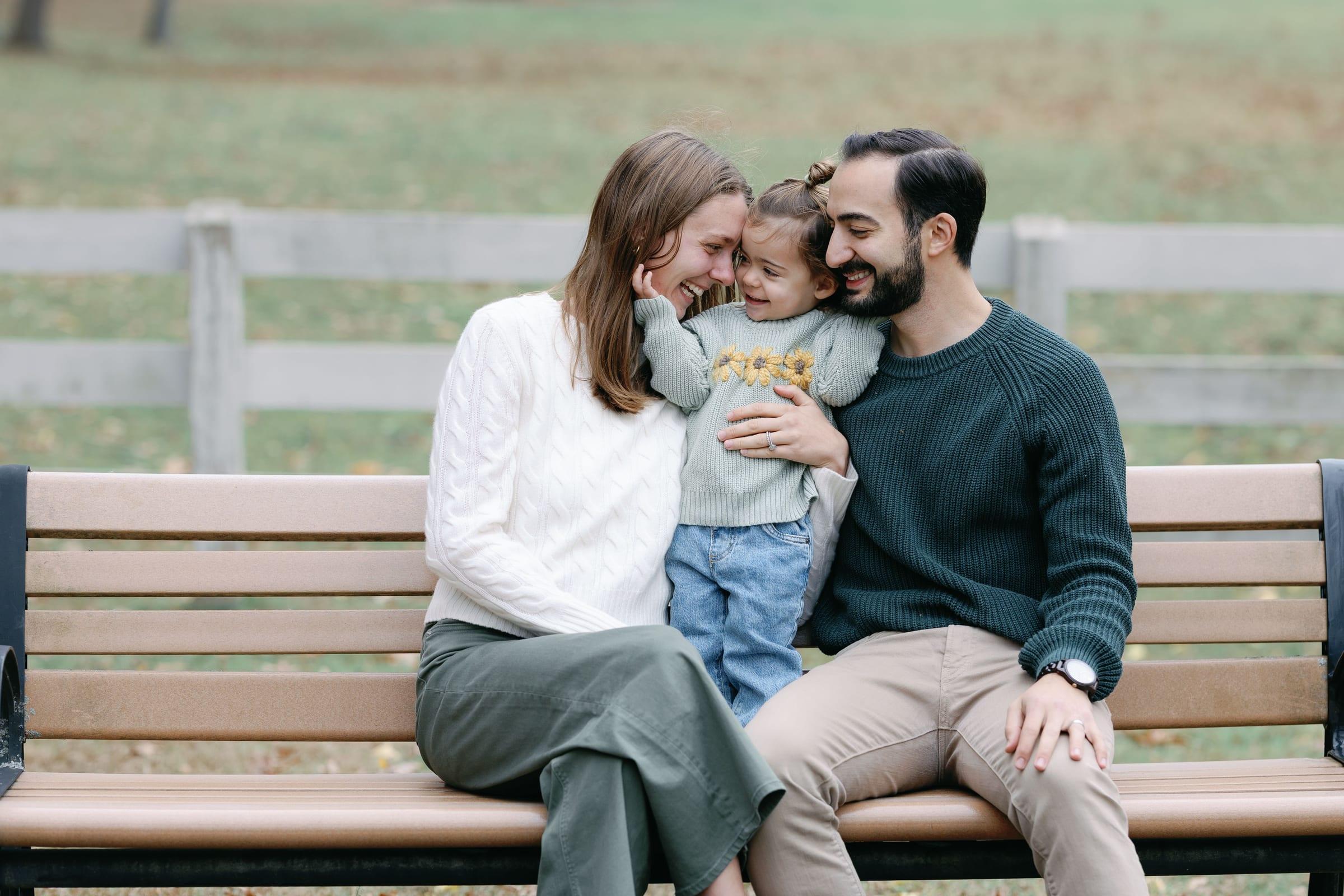 A mother, father, and toddler sit close together on a park bench, smiling at one another.