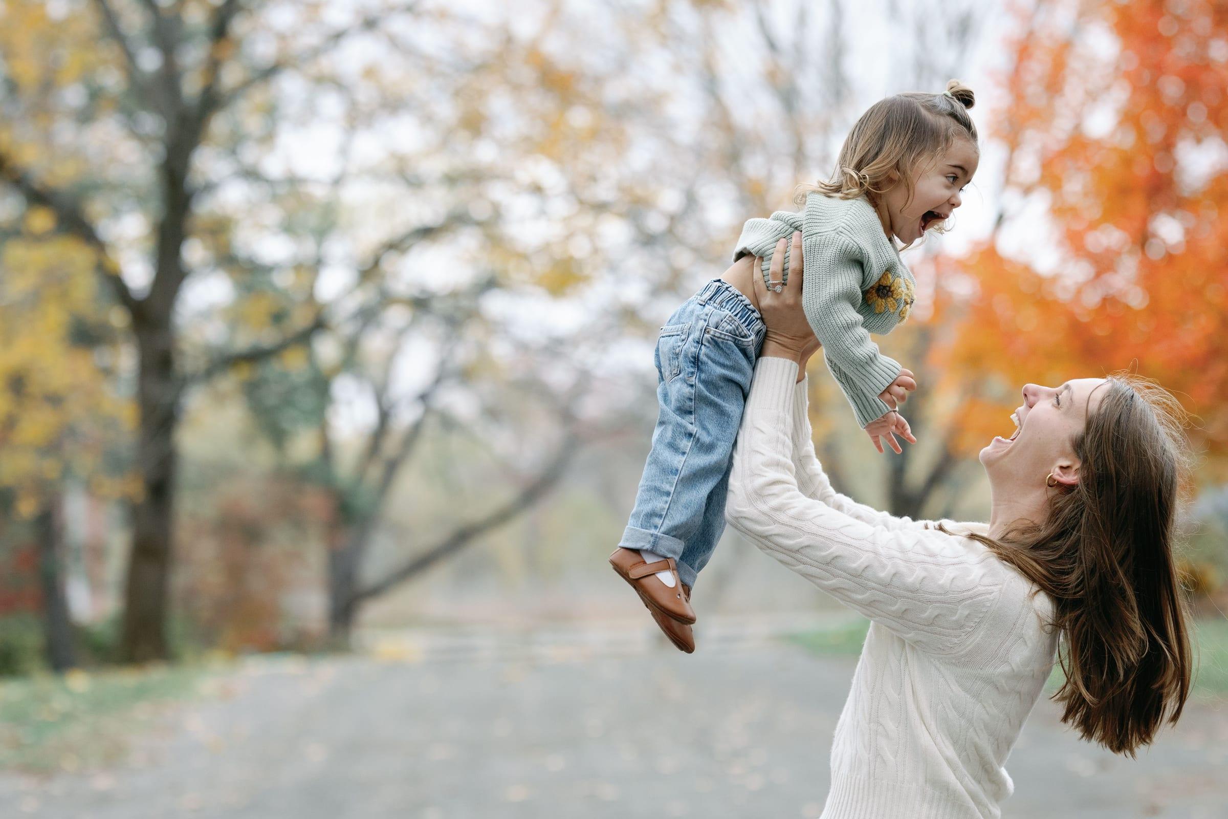 A mother lifts her laughing toddler high in the air in an autumn park.